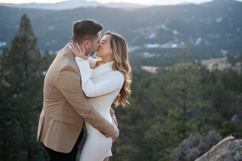 engaged couple kissing in a mountain setting wearing a white dress and tan jacket